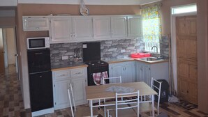 A compact kitchen with white cabinets, a microwave, a stove, and a sink. There is a dining table with chairs and a gray sofa. The floor has a checkered pattern.