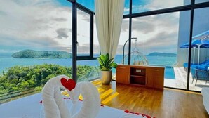 Una habitación con un gran ventanal, una cama con ropa blanca, un escritorio de madera y vistas a una masa de agua y vegetación.
