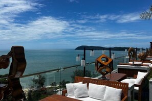 Una terraza frente al mar con mobiliario de madera, barandilla de cristal y vista al océano.