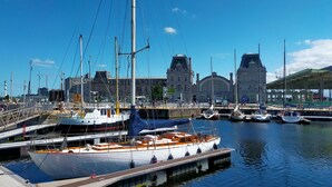 Un port de plaisance où sont amarrés plusieurs bateaux, un grand bâtiment au style architectural particulier en arrière-plan et un ciel bleu dégagé.