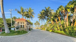 A paved pathway leading to a building with a red-tiled roof, surrounded by lush greenery and palm trees.