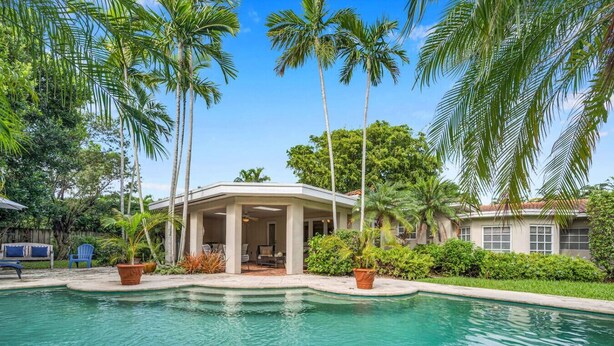 A pool with a covered patio area, surrounded by palm trees and a house.