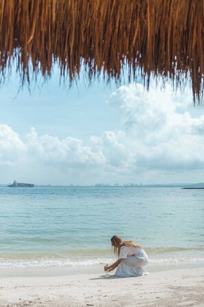 A woman in a white dress sitting on a beach near the water.