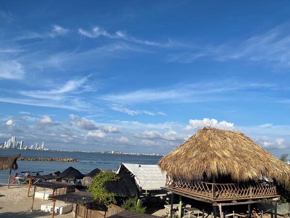 A beach with thatched-roof huts and a view of a city skyline across the water.