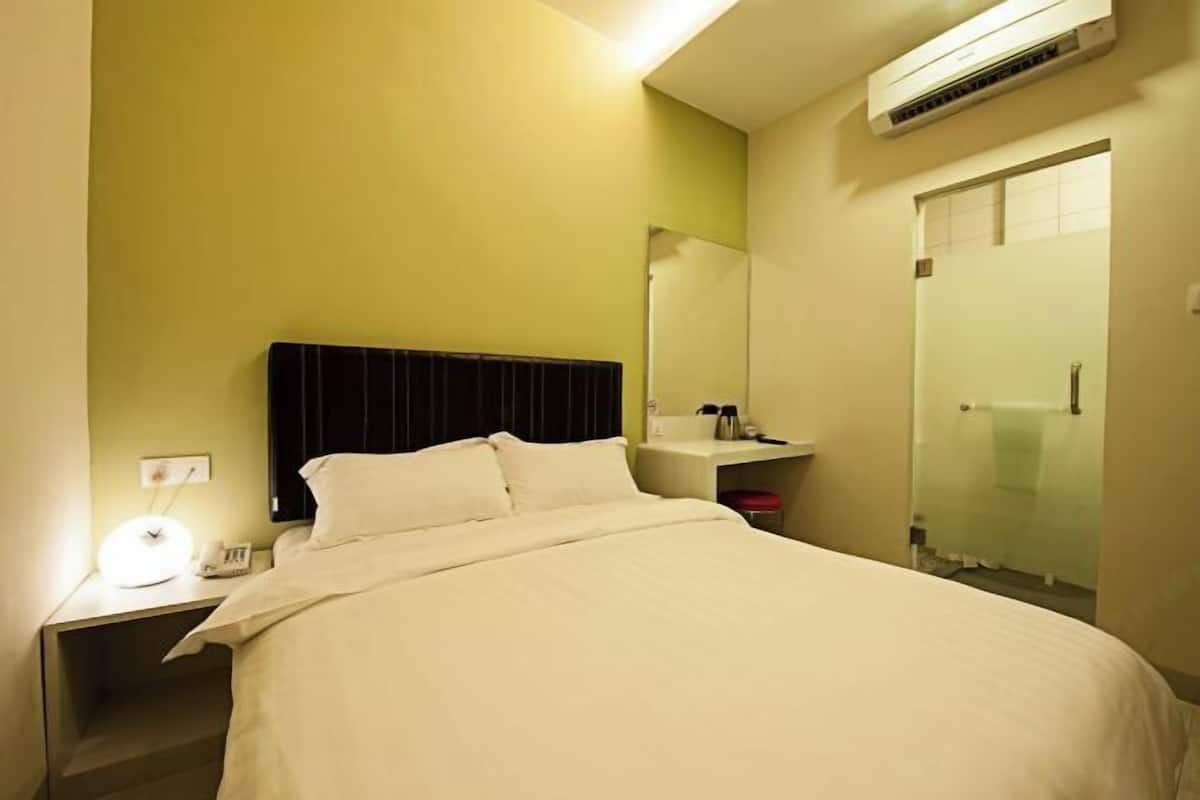 A hotel room with a bed, bedside table, lamp, and a bathroom with a glass shower enclosure.