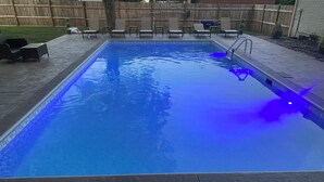 A rectangular swimming pool with a tiled edge, surrounded by a patio area with lounge chairs and a wooden fence in the background.