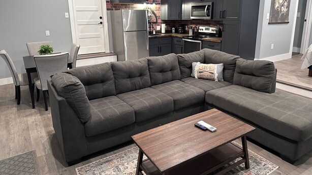 A modern living room with a sectional sofa, a wooden coffee table, and a kitchen area with grey cabinets and a stainless steel refrigerator.