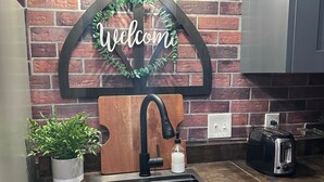 A kitchen with a brick wall, a welcome sign, a sink, a toaster, and a potted plant.