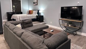 A modern living room with a grey sofa set, a flat-screen TV on a curved stand, a bed with a white headboard, and a small table with a remote control.