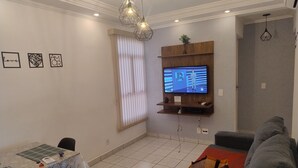 A living room with a TV mounted on a wooden shelf, a dining table with a patterned tablecloth, and a grey sofa with colorful cushions. - Easy to locate apartment with air conditioning (Itupeva)