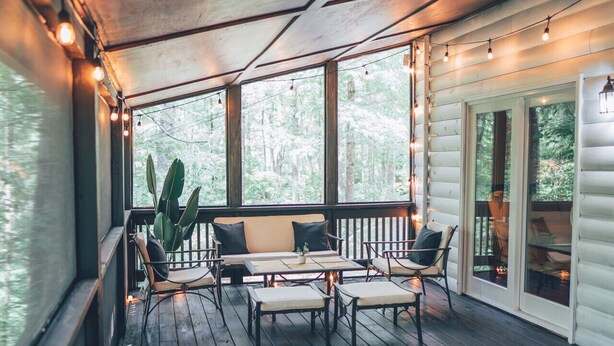 Una terraza techada al aire libre con suelo de madera, un sofá y sillas, rodeada de amplios ventanales y luces colgantes.