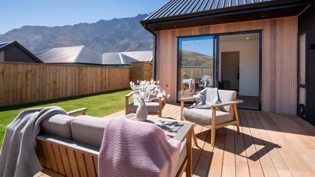 A modern outdoor deck with a wooden floor, a sofa, a chair, and a table. There is a wooden fence in the background and a mountain range in the distance. - Remarkable stay in Queenstown (Jacks Point)
