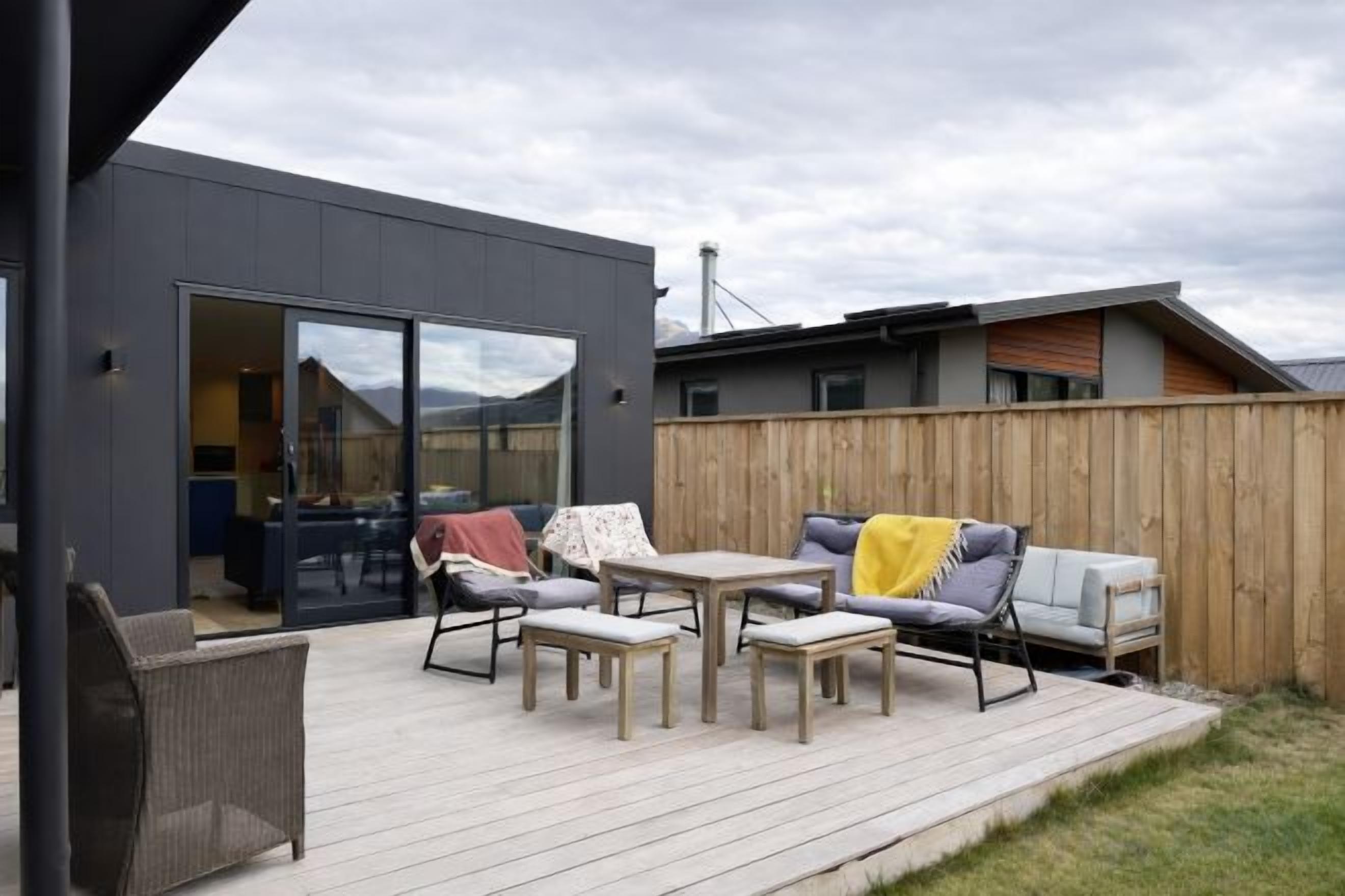 Un patio exterior con un comedor y un sofá, una terraza de madera y un edificio moderno con puerta de vidrio corredera.