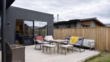 Un patio exterior con un comedor y un sofá, una terraza de madera y un edificio moderno con puerta de vidrio corredera.