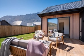A wooden deck with outdoor furniture, a glass door leading to a room, and a mountain backdrop.