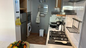 Eine kompakte Küche mit Herd, Spüle und einer kleinen Arbeitsfläche. Eine Waschmaschine und ein Wäscheständer sind vorhanden. Auf der Arbeitsfläche steht eine Schüssel mit Obst.