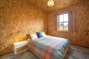 A bedroom with a wooden wall, a bed with a colorful blanket, a nightstand, a wall-mounted lamp, and a window.
