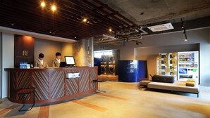 Un espace d’accueil d’hôtel comprenant un comptoir de réception, deux réceptionnistes, un coin salon et un distributeur automatique.
