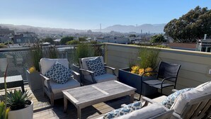 A rooftop patio with outdoor furniture, potted plants, and a view of the cityscape and bridge in the distance. - Sunny 3 Bedroom -Best San Francisco Neighborhood-Roofdeck w. Epic Views (San Francisco)