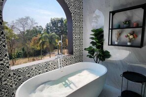 Une salle de bain avec une baignoire sur pieds, une fenêtre donnant sur la verdure et une étagère ornée d’objets décoratifs.