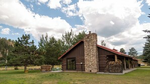 A log cabin with a stone chimney, surrounded by a grassy field and trees.
