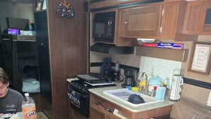 A compact RV kitchen with a stove, microwave, and sink. A person is seated at a table with snacks and a drink. - Cozy RV in peaceful Sandia with WiFi, AC,Lake Corpus Chrisit Access nearby (Sandia)