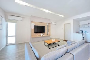 A modern living room with a sofa, a coffee table, a TV mounted on the wall, and a kitchen area with a washing machine. - Restful Respite Tanjung Aru Inifnity Pool 2BR (Kota Kinabalu)
