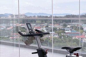 A stationary bike with a digital display, positioned in a room with large windows overlooking a residential area. - Restful Respite Tanjung Aru Inifnity Pool 2BR (Kota Kinabalu)