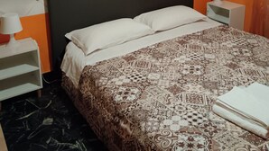 Ein ordentlich bezogenes Bett mit einem gemusterten Überwurf, ein Kopfteil mit einer gepolsterten Optik und ein weißer Nachttisch mit einer Lampe.