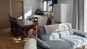 A modern living room with a grey sofa, wooden dining table, and chairs. There is a kitchen area with a refrigerator, stove, and window.