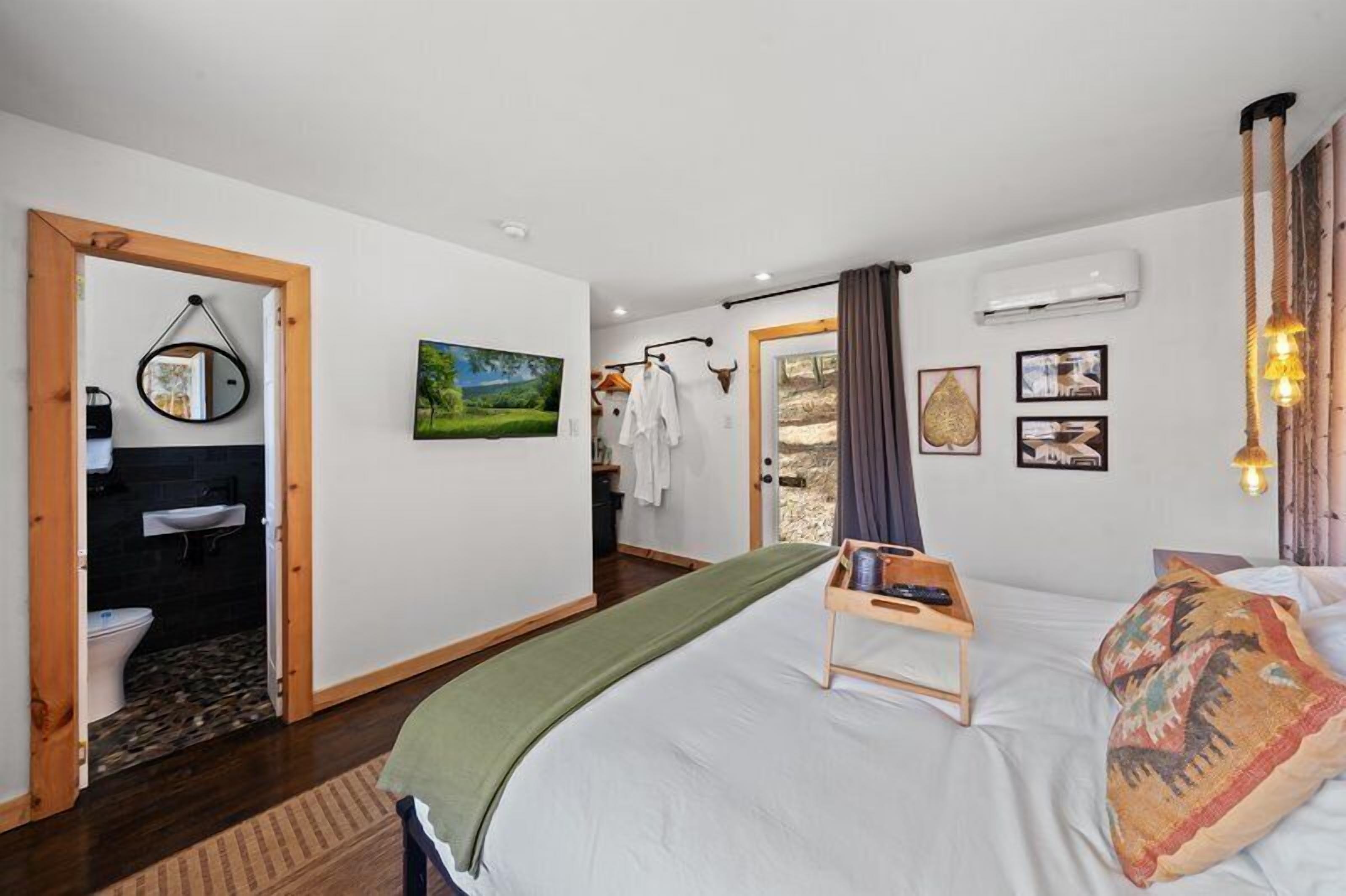 Un dormitorio con cama, mesita de noche, espejo colgado, cuadro en la pared y baño visible a través de una puerta abierta.