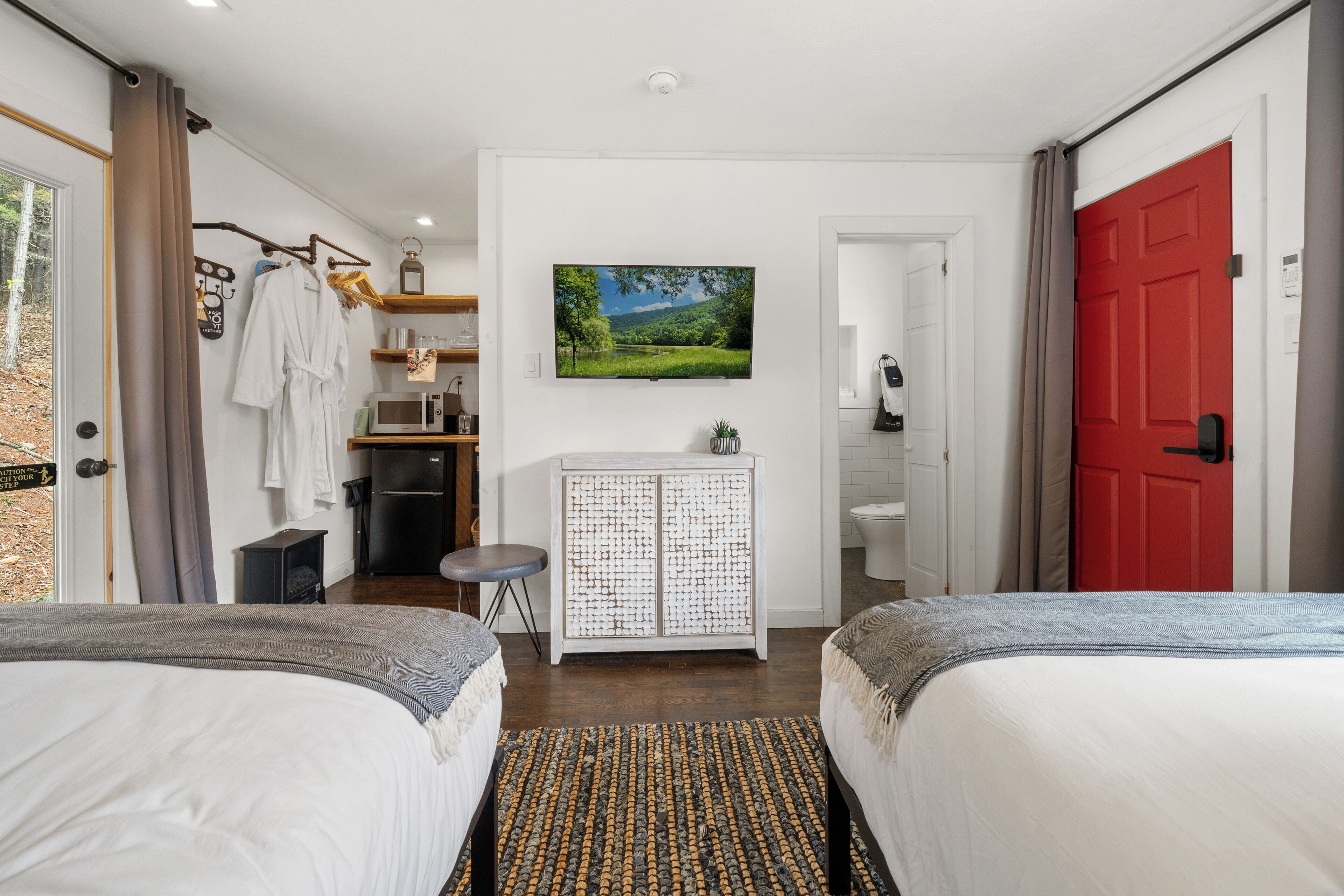 Habitación de hotel con dos camas, una kitchenette pequeña, un baño y un ventanal con vistas a árboles.
