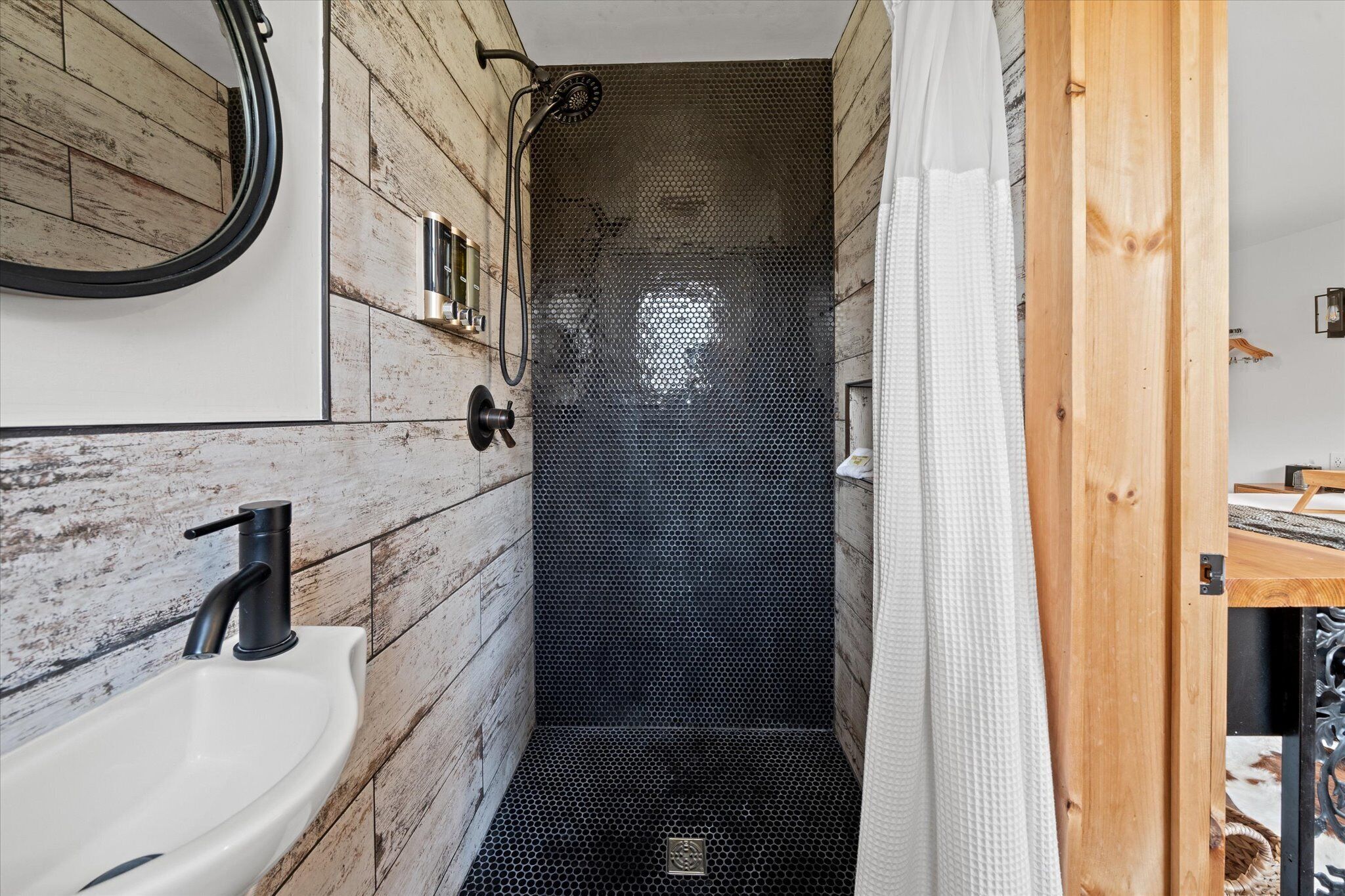 A narrow bathroom with a wooden wall, a white sink, and a shower with a mesh door.