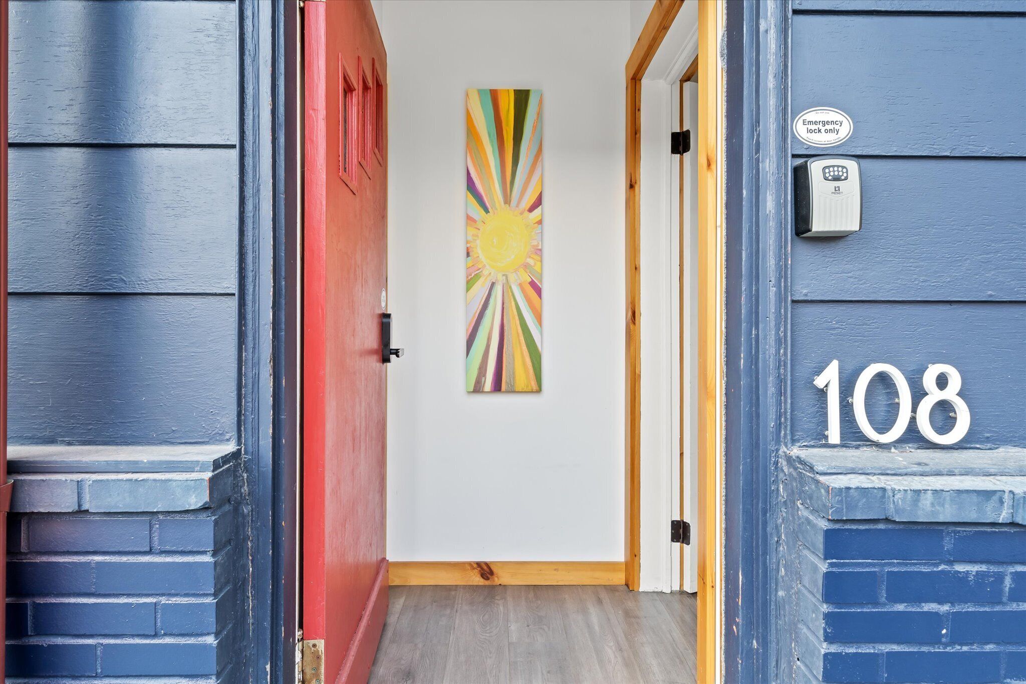 A hallway with a red door, a colorful sunburst painting, and a blue exterior wall with the number 108.