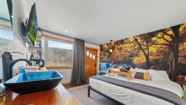 Un dormitorio con cama, ventana grande, televisor montado en la pared, lavamanos y vistas a un paisaje montañoso.