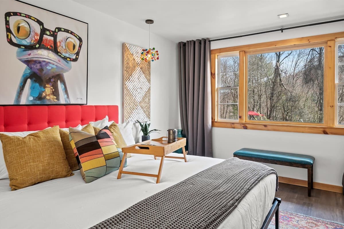 A bedroom with a red upholstered headboard, a large window with a view of trees, a wooden bedside table with a plant, and a bench.