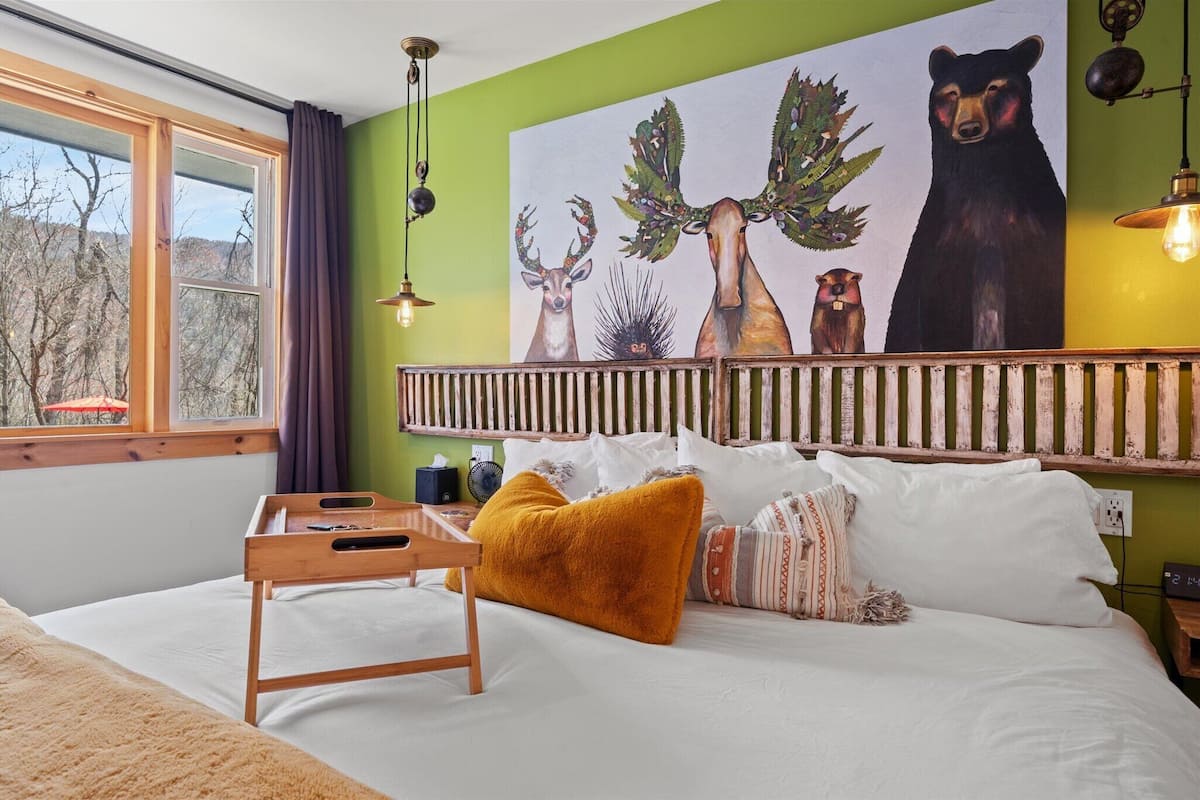 A bedroom with a bed, a wooden bedside table, a nightstand, a wall-mounted shelf, a window with curtains, and a painting of deer and a bear.
