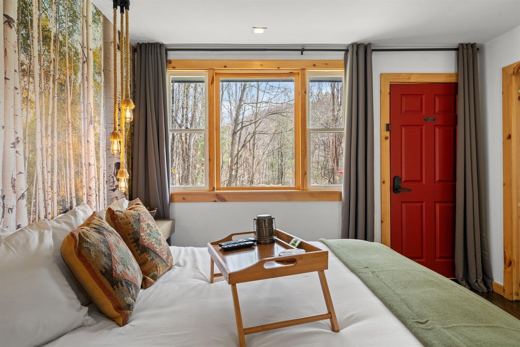 A bedroom with a large bed, a wooden bedside table with a tray, a red door, and a window with a forest view.