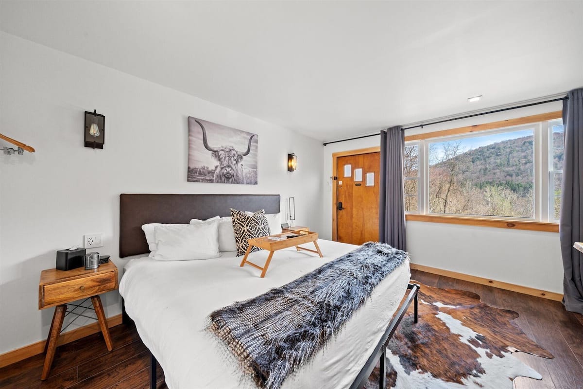 A bedroom with a large bed, a wooden nightstand, a bedside table, a wall-mounted light, a framed picture, a window with curtains, and a view of a mountainous landscape.