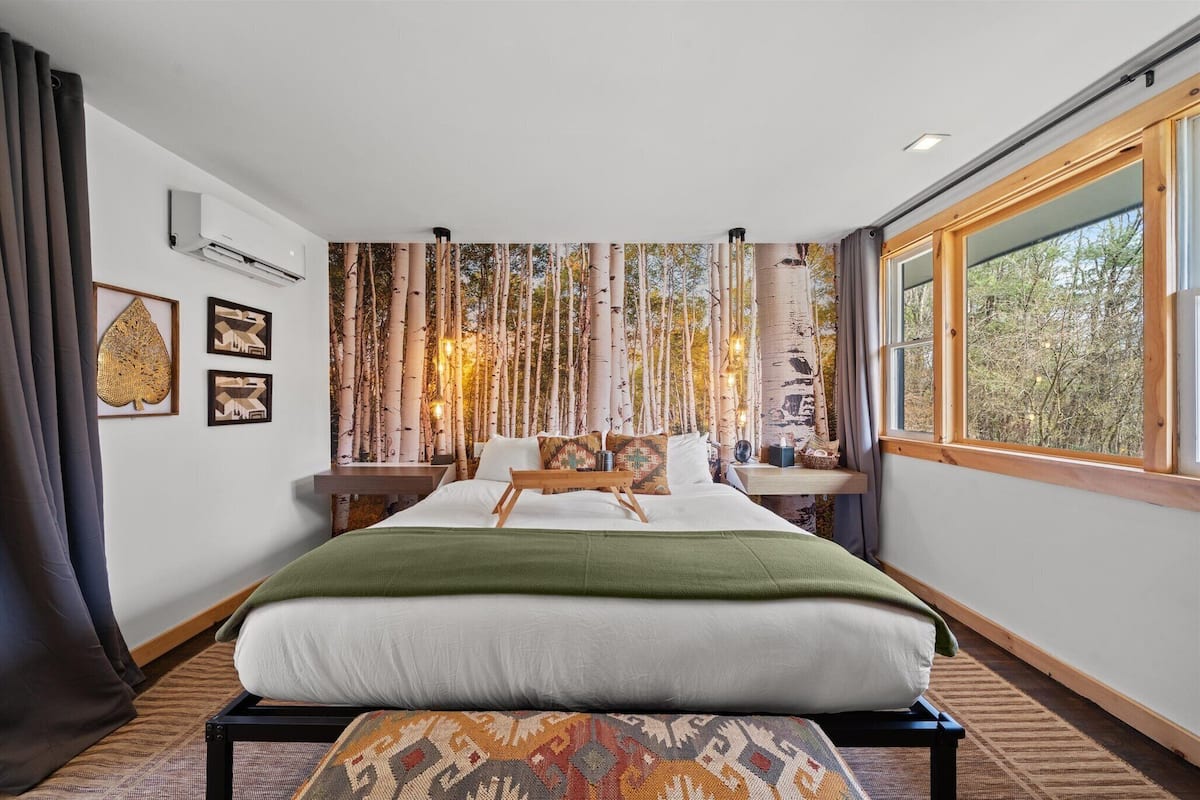 A modern bedroom with a large bed, a view of trees through the window, and a wall-mounted air conditioner.