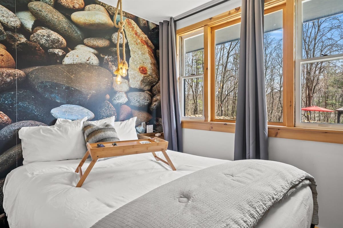 A bedroom with a large bed, a wooden bedside table, a wall mural of rocks, and a window with a view of trees.
