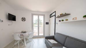 A modern living room with a grey sofa, a dining table with chairs, a flat-screen TV, a wall clock, and a sliding glass door leading to an outdoor area.