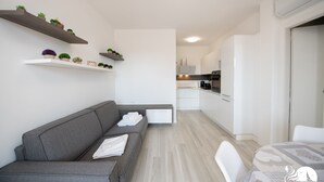 A modern living room with a grey sofa, white chairs, and a dining table. There are shelves with plants and a kitchen in the background. - Villa Liliana - Apt 06 (Sirmione)