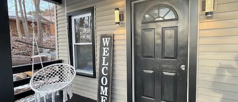 A porch with a swing chair, a 'WELCOME' sign, and a dark door.