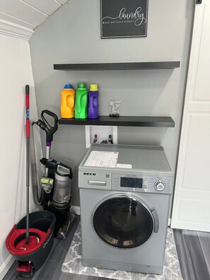 A laundry area with a washing machine, a mop, cleaning supplies, and a shelf with a sign. - Lakefront Home w/ Dock & Fire Pits (Northville)