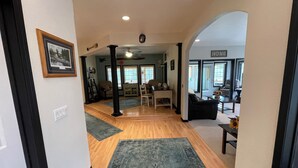 A spacious living area with a wooden floor, a black sofa, a coffee table, and a dining table with chairs. There are multiple windows providing natural light and a view of the outside. - “Wake Up by the Lake — Private Alaska Stay Near Hiking & ATV Trails” (seward)