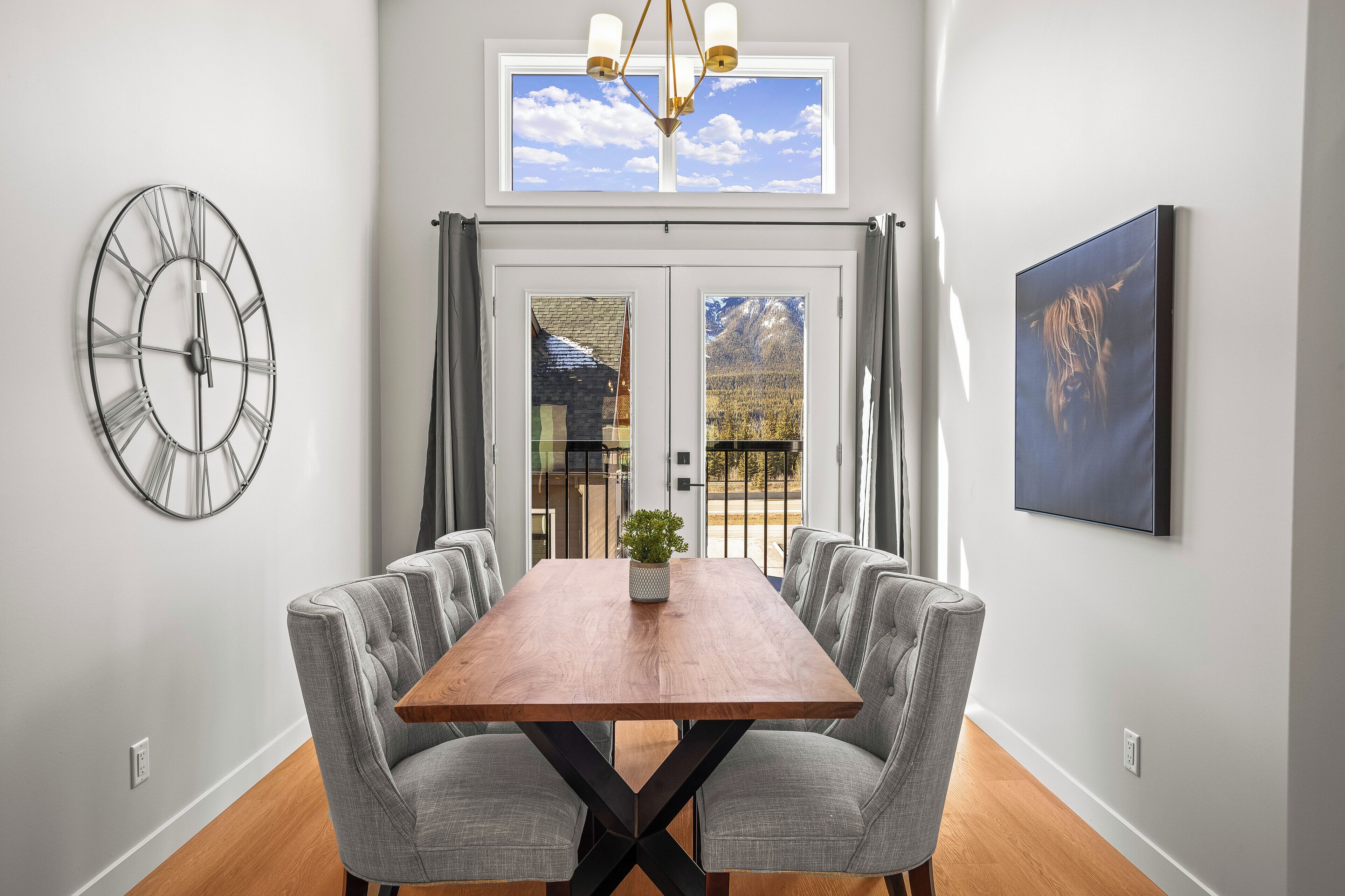 Un coin repas avec une table en bois, des chaises à revêtement gris, une horloge murale, une fenêtre donnant sur les montagnes et un téléviseur fixé au mur.