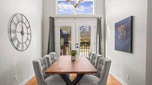 A dining area with a wooden table, grey upholstered chairs, a wall clock, a window with a view of mountains, and a mounted television.