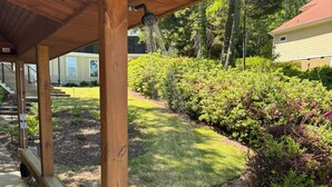 Una terraza de madera con borde de piedra, un espacio cubierto con techo de madera y un jardín ajardinado con arbustos variados y césped.