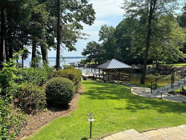 A gazebo with a metal roof overlooking a lake, surrounded by lush greenery and a well-maintained lawn.